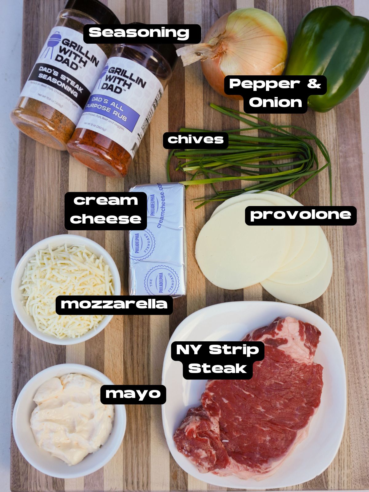 ingredient list of the cheesesteak dip. Bottles of seasoning, yellow onion, green bell pepper, chives, shredded mozzarella cheese, cream cheese, provolone cheese slices, a bowl of mayo and a plated raw NY strip.