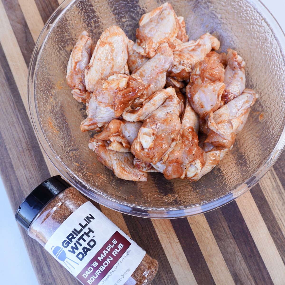 seasoned raw chicken wings in a clear bowl with a bottle of maple bourbon seasoning next to it.
